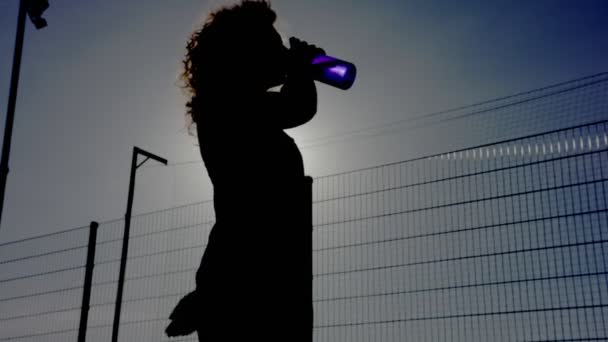 Silhouette di una bella donna, beve acqua da una bottiglia d'acqua blu, traslucida, sullo sfondo del sole. video in 4K
. - Filmati, video