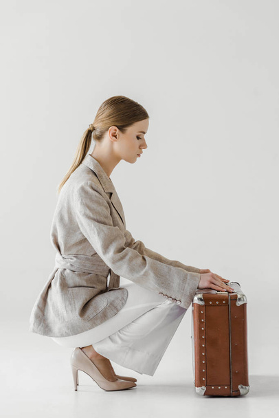 side view of stylish young female traveler in linen jacket opening vintage suitcase isolated on grey background  - Фото, изображение