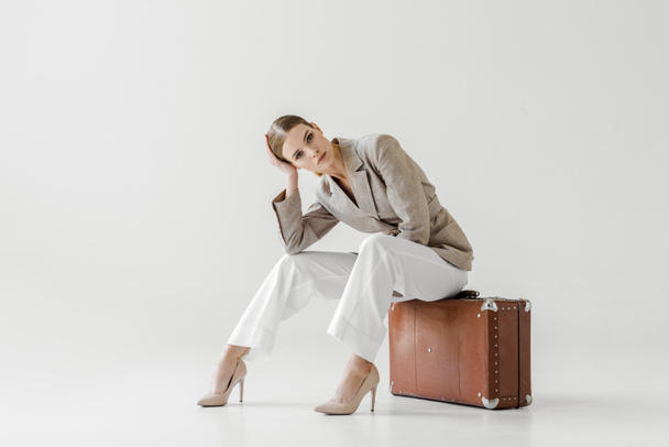 young stylish female model in linen jacket sitting on vintage suitcase and looking at camera isolated on grey background  - Φωτογραφία, εικόνα