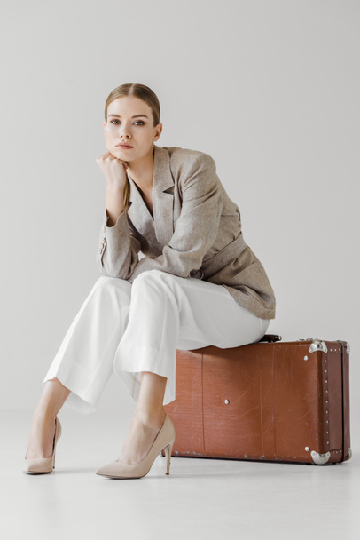stylish young female model in linen jacket with hand on chin sitting on vintage suitcase isolated on grey background  - Valokuva, kuva