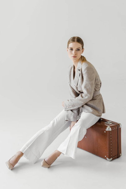 attractive stylish woman in linen jacket sitting on vintage suitcase isolated on grey background  - Foto, Imagen