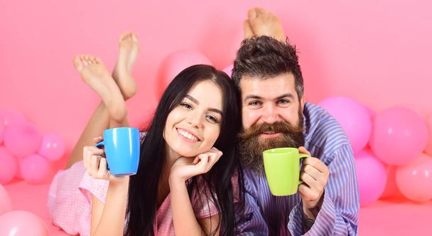 Man and woman on smiling faces lay, pink background. Couple in love drink coffee in bed. Couple relax in morning with coffee. Family tradition concept. Man and woman in domestic clothes, pajamas - Фото, зображення