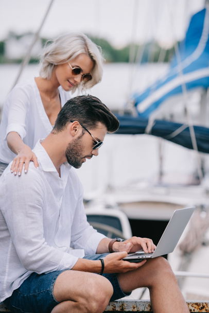 side view of smiling girl looking at handsome man in sunglasses using laptop on yacht - Fotó, kép