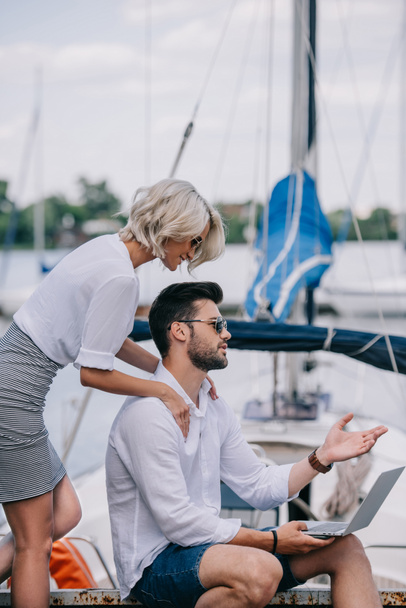 smiling girl looking at handsome man in sunglasses using laptop on yacht - Foto, Imagem