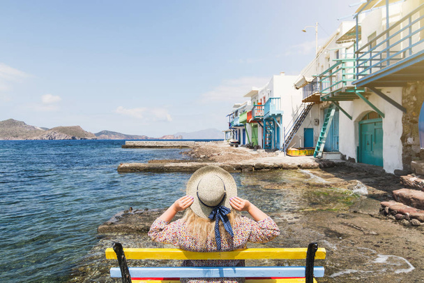 Молодая блондинка сидит на скамейке с видом на цветные старые дома в рыбацком городке Клима на острове Милош, Греция. Солнечный день, широкоугольный объектив
 - Фото, изображение