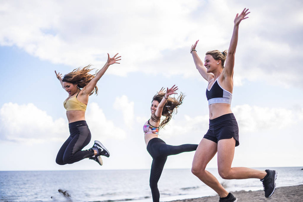 three beautiful  Caucasian girls jumping on the beach doing fitness workout. outdoor leisure sport activity for group of females people with ocean in background - Photo, Image