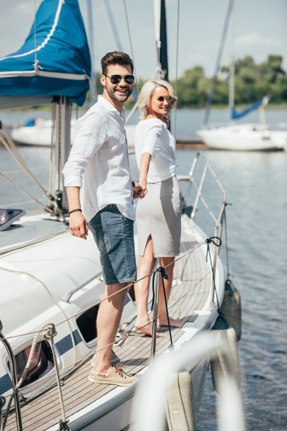 サングラス手を繋いでいると、ヨットの上でカメラに笑顔で美しい幸せな若いカップル - 写真・画像