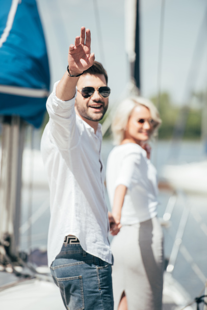 giovane uomo in occhiali da sole sorridente alla fotocamera e agitando la mano mentre si tiene per mano con bella ragazza su yacht
  - Foto, immagini