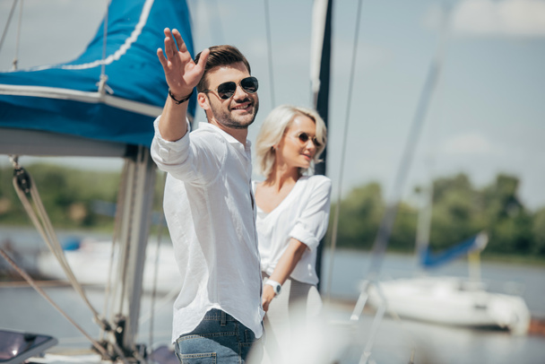 happy young man in sunglasses smiling at camera and waving hand while holding hands with beautiful girl on yacht  - Photo, Image