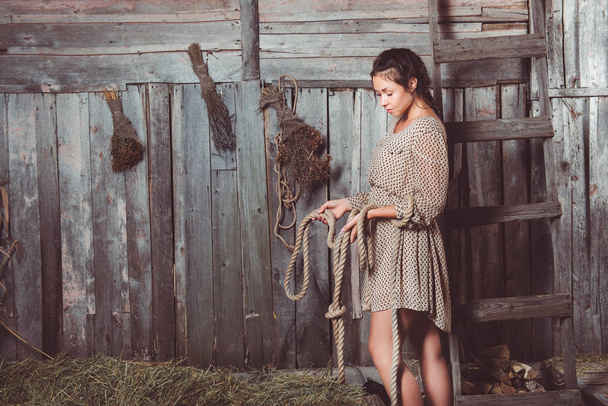 Young girl in the barn holding rope in her hands and looking at it, ladder and brooms on her background - Foto, Bild