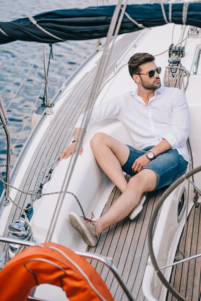 handsome young man in sunglasses looking away while sitting on yacht - Photo, Image