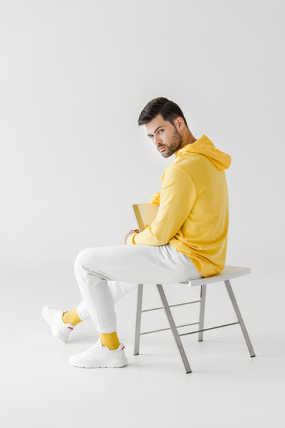 attractive young man in yellow hoodie sitting on flipped chair on white and looking at camera - Φωτογραφία, εικόνα