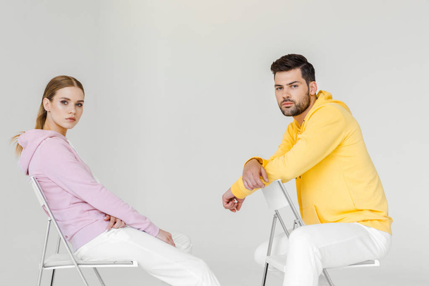 vista lateral de los jóvenes modelos masculinos y femeninos en sudaderas con capucha rosa y amarilla sentados en sillas y mirando a la cámara en blanco
 - Foto, imagen