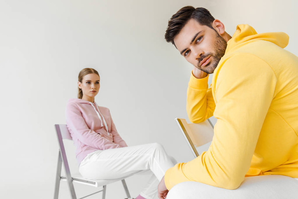 stylish young male and female models in pink and yellow hoodies sitting on chairs isolated on white - Foto, afbeelding