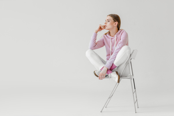 thoughtful young woman in pink hoodie sitting on chair on white and looking away - 写真・画像