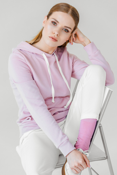 sensuele jongedame in roze hoodie zittend op een stoel op wit en camera kijken - Foto, afbeelding