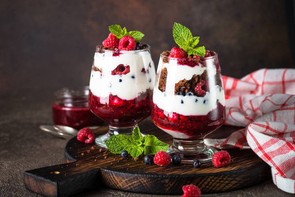 Слоистый десерт со сметаной, шоколадным бисквитом, джемом и свежими ягодами в стеклянной банке
. - Фото, изображение