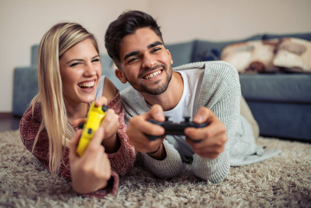 Χαρούμενο ζευγάρι παίζουν βιντεοπαιχνίδια ενώ ξαπλωμένη σε ένα χαλί. - Φωτογραφία, εικόνα