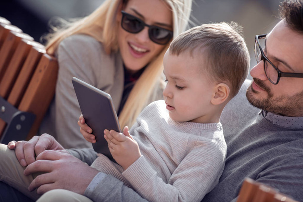 Onnellinen perhe ja digitaaliteknologia.Nuoret vanhemmat ja pieni lapsi käyttävät tablettia istuessaan kaupungin puistossa
. - Valokuva, kuva