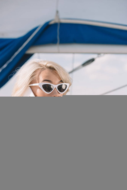 Schockierte junge Frau in Sonnenbrille und Badehose spricht auf Smartphone und liegt auf Jacht  - Foto, Bild