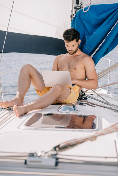 focused shirtless man in swim trunks using laptop on yacht - Foto, Bild