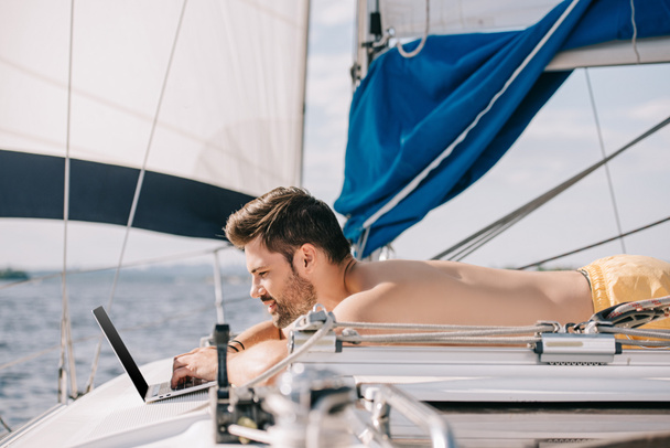 vue latérale d'un homme torse nu souriant utilisant un ordinateur portable sur un yacht
  - Photo, image