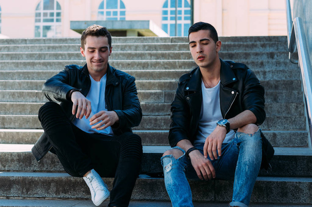 two guys in black jackets, white T-shirts, jeans and white shoes are sitting on the steps. on the hands of a watch. socializing, meeting friends. in the city near the railings - Φωτογραφία, εικόνα