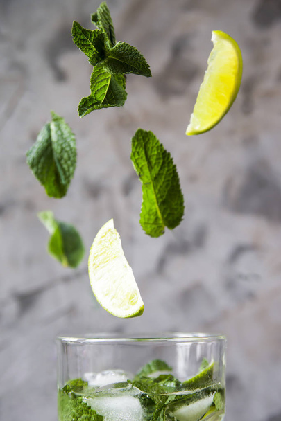 Des feuilles de menthe et de citron vert volent dans un verre avec du mojito avec de la glace sur un fond gris. Lévitation vol objets magiques. Boisson d'été
 - Photo, image