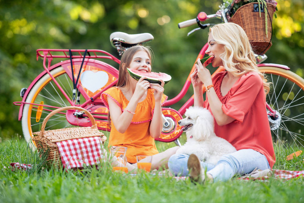 Kaunis nuori äiti ja hänen tyttärensä istuvat nurmikolla ja syövät vesimelonia.
. - Valokuva, kuva