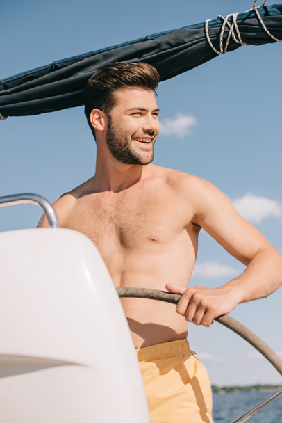 smiling shirtless muscular man in swim trunks steering yacht  - Fotó, kép