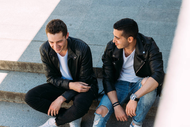 two guys in black jackets, jeans and white shoes, in white T-shirts are sitting on the steps. on the hands of a watch. socializing, meeting friends. - Foto, Imagen