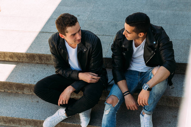 two guys in black jackets, jeans and white shoes, in white T-shirts are sitting on the steps. on the hands of a watch. socializing, meeting friends. - Foto, Bild