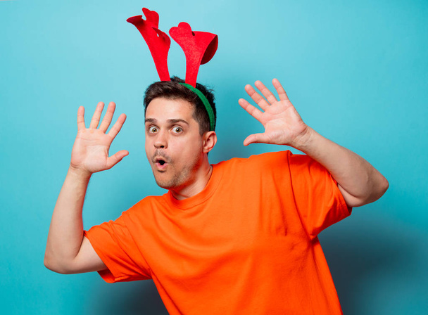 Joven hombre guapo en camiseta naranja con cuernos de ciervo de Navidad. Imagen del estudio sobre fondo azul
 - Foto, Imagen