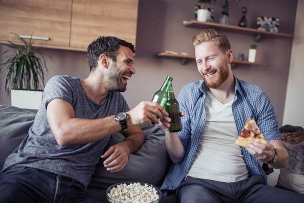 Amigos comendo pipocas, bebendo cerveja e comendo pizza juntos e assistindo jogo de futebol no sofá em casa.
. - Foto, Imagem