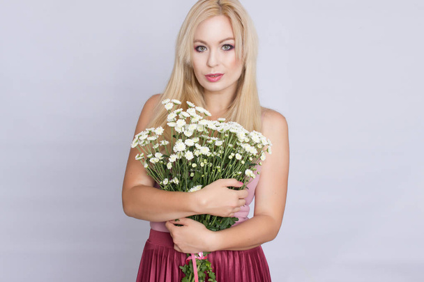 Portrait of a happy blonde woman in pink attire holding a bouquet of white wildflowers in hands - Foto, afbeelding