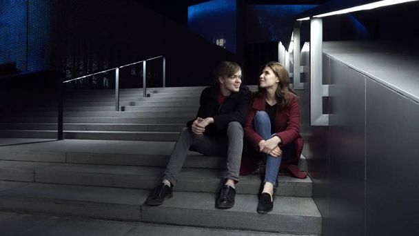 Young couple sitting on stairs at night. Beautiful night for a walk around the city - Zdjęcie, obraz
