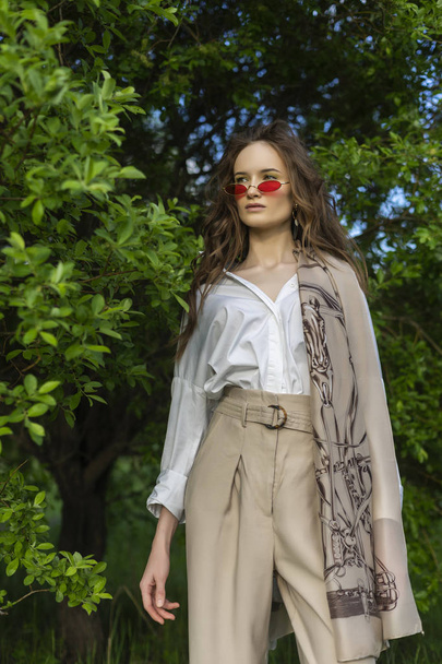 Beautiful stylish young girl wearing a trendy white shirt, beige trousers, pareo and red sunglasses poses outdoors against a background of green leaves. Commercial and advertisement design. Copy space - Fotoğraf, Görsel