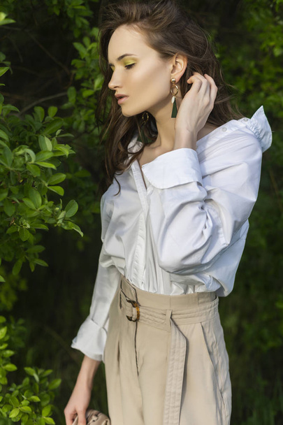Beautiful stylish young girl wearing a trendy white shirt, beige trousers and a pareo poses outdoors against a background of green leaves. Nude makeup. Commercial and advertisement design. Copy space. - Foto, afbeelding