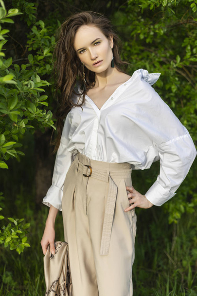 Beautiful stylish young girl wearing a trendy white shirt, beige trousers and a pareo poses outdoors against a background of green leaves. Nude makeup. Commercial and advertisement design. Copy space. - Φωτογραφία, εικόνα