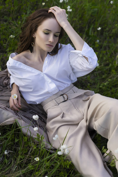 Όμορφο κομψό νεαρή κοπέλα φορώντας ένα μοντέρνο λευκό πουκάμισο, μπεζ παντελόνι γυμναστικής και ένα παρεό βρίσκεται χαλαρώνοντας στο πράσινο γρασίδι ανάμεσα στα αγριολούλουδα. Σχεδιασμός εμπορικής και διαφήμιση. Χώρο αντίγραφο. - Φωτογραφία, εικόνα