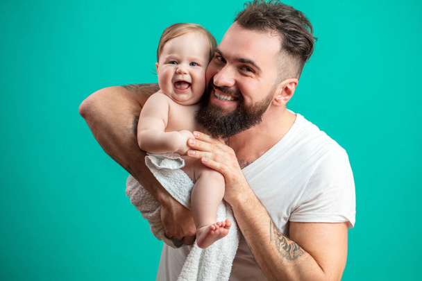 Caucasian handsome guy with stylish beard hugging and playing with his beautiful infant daughter, looking at camera, over blue background, expressing care and tenderness. - Photo, Image