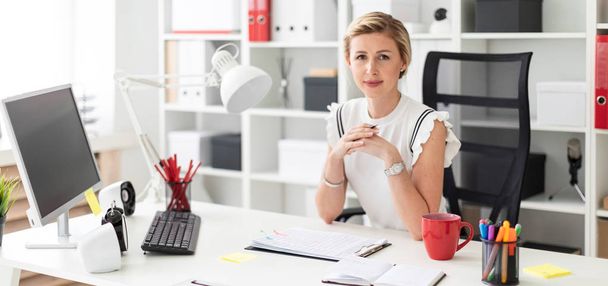 Una joven rubia se sienta en un escritorio de computadora en la oficina y sostiene un lápiz en su mano
. - Foto, Imagen