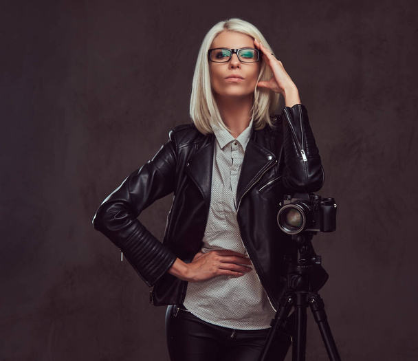 Intelligente fotografa bionda in abiti alla moda in posa mentre si appoggia su una fotocamera professionale con un treppiede in uno studio, guarda la fotocamera
. - Foto, immagini