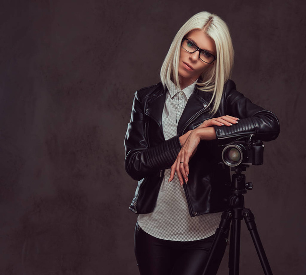 Mujer rubia inteligente fotógrafa en ropa de moda posando mientras se apoya en una cámara profesional con un trípode en un estudio, mira a la cámara
. - Foto, Imagen