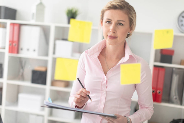 A young blonde girl stands in the office next to a transparent board with stickers and holds documents and a pencil in her hands. - Photo, Image
