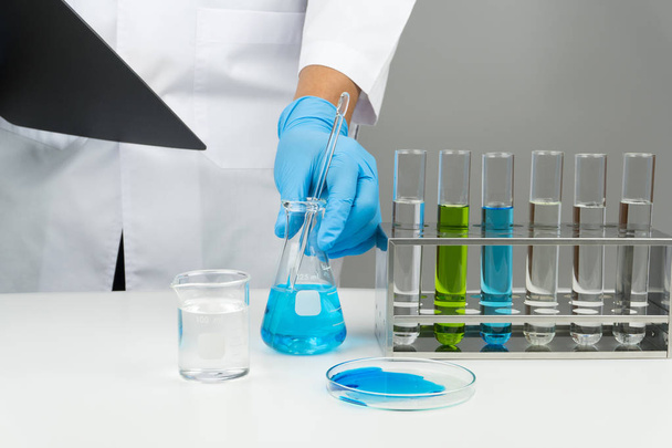 ガラス攪拌棒、ラック、時計ガラス テーブルの上のビーカー、試験管とクリップボードと青液フラスコを保持している科学者の研究室シーン - 写真・画像