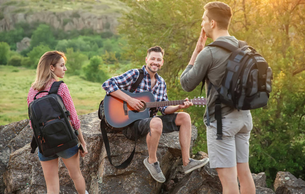 Sırt çantası için arkadaşları wilderness içinde gitar çalmak olan genç adam. Kamp sezon - Fotoğraf, Görsel
