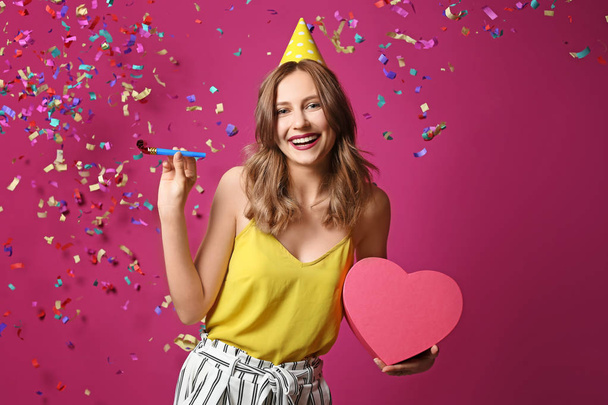 Young woman with birthday gift and party blower on color background - Foto, Imagen