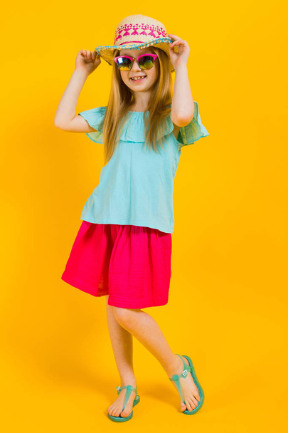 Girl with red hair on a yellow background. A charming girl in bright summer clothes holds her hand for her hat. - Foto, imagen