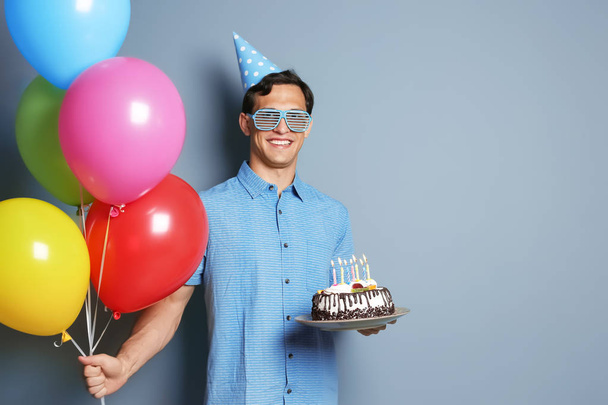 Young man with birthday cake and bright balloons on color background - Φωτογραφία, εικόνα